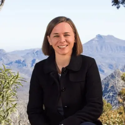 Professor Lisa Harvey-Smith is sitting outdoors, smiling at the camera. There are mountains in the background, she is a fair-skinned woman with shoulder length brown hair. She is wearing dark clothing.